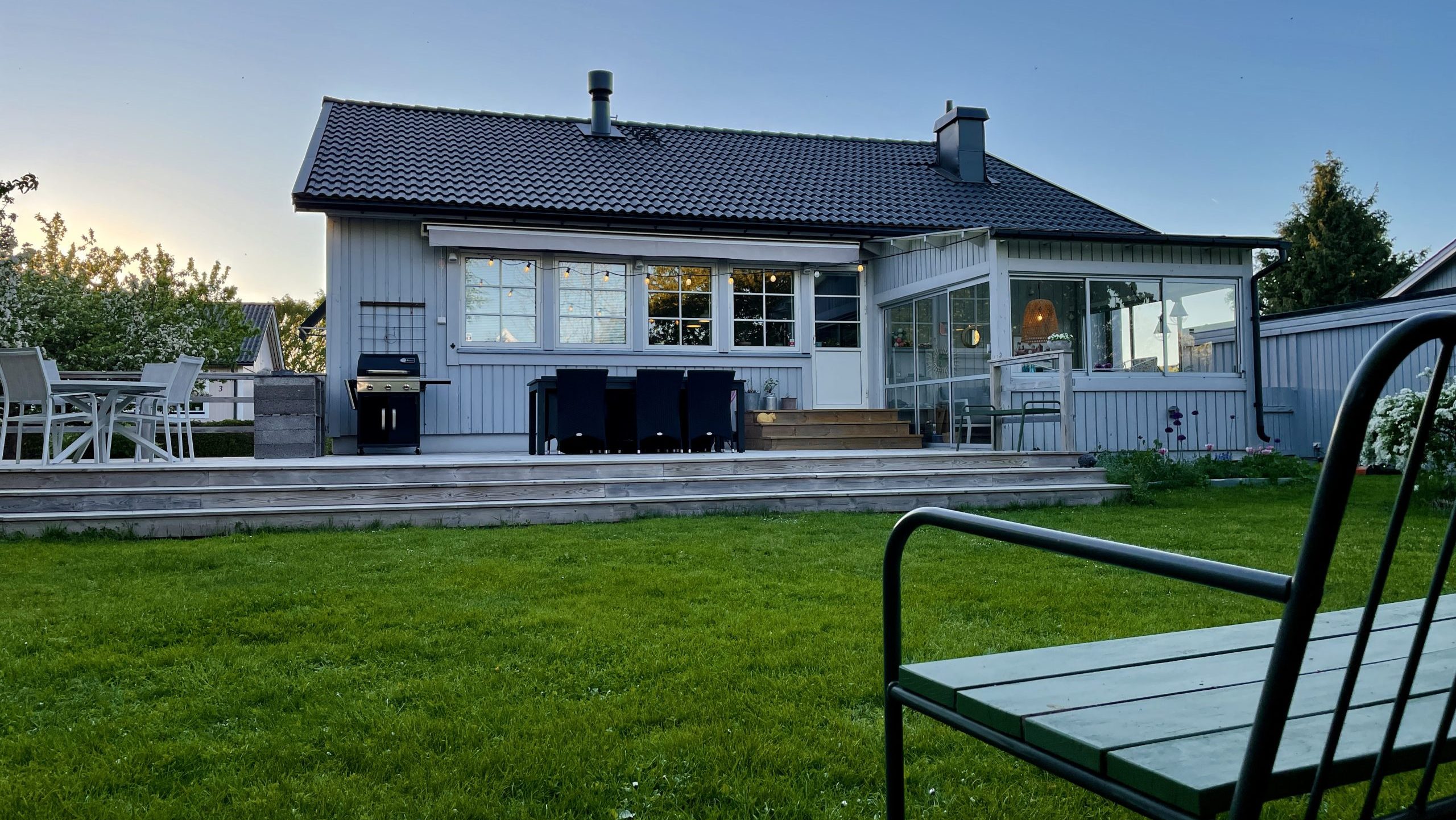 Grått hus med villaträdgård och bänk i förgrunden.